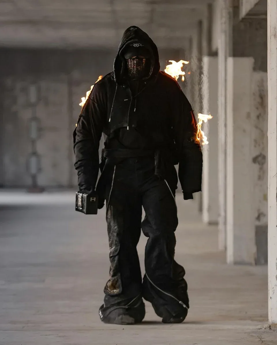 The professional stuntman wore a black hooded outfit and carried a box bag while flames rose behind him.