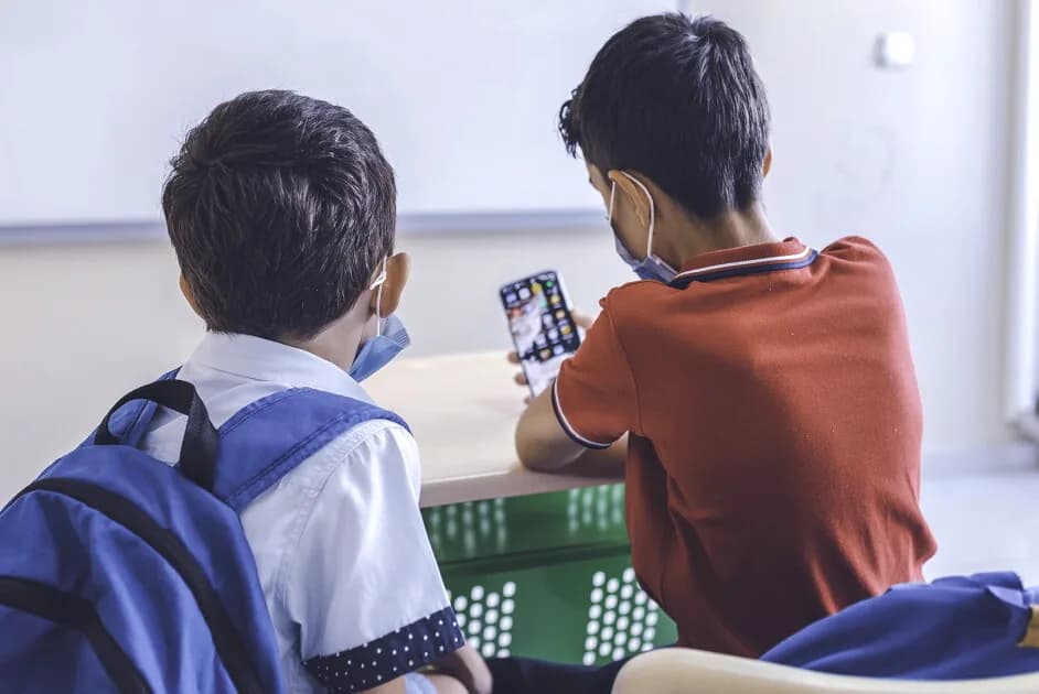 Close up rear view of two schoolboys wearing protective n95 face mask looking, playing games, working for online education with smartphone in a classroom during COVID-19 outbreak