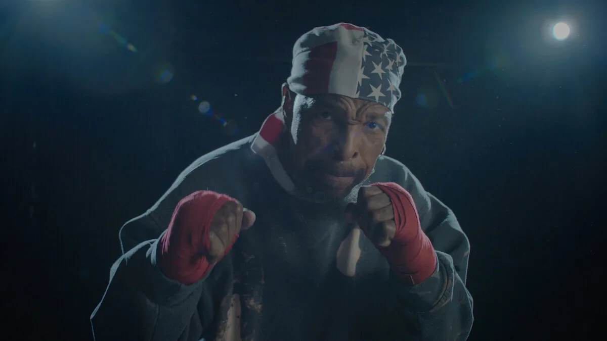 A male boxer wearing red hand wraps and a USA flag bandana poses in a fighting stance under dramatic lighting in a dark gym or training environment.