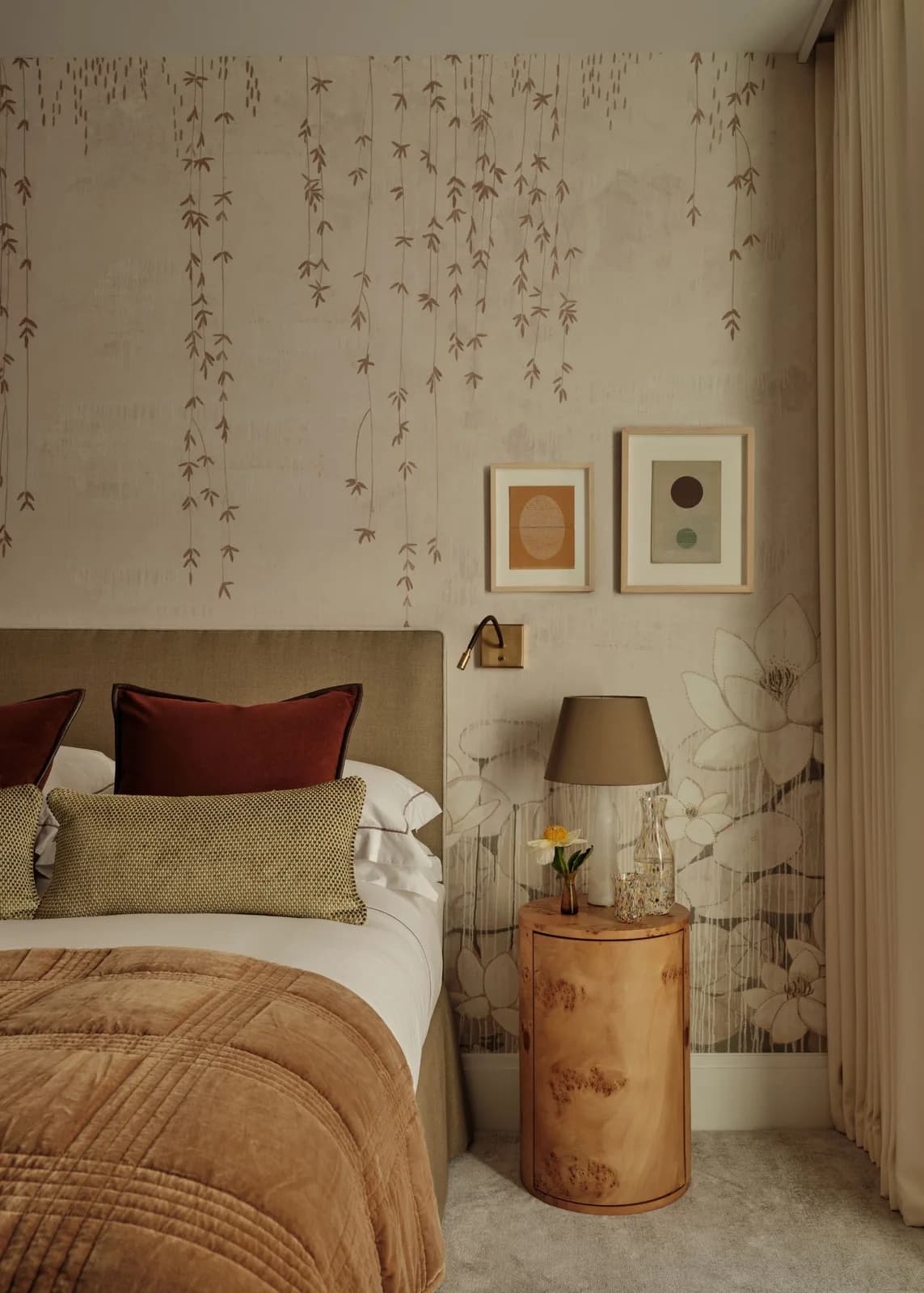 A bedroom with a beige floral patterned wallpaper, beige carpeted floor, a taupe upholstered headboard with layered neutral bedding and a burl wood nightstand.