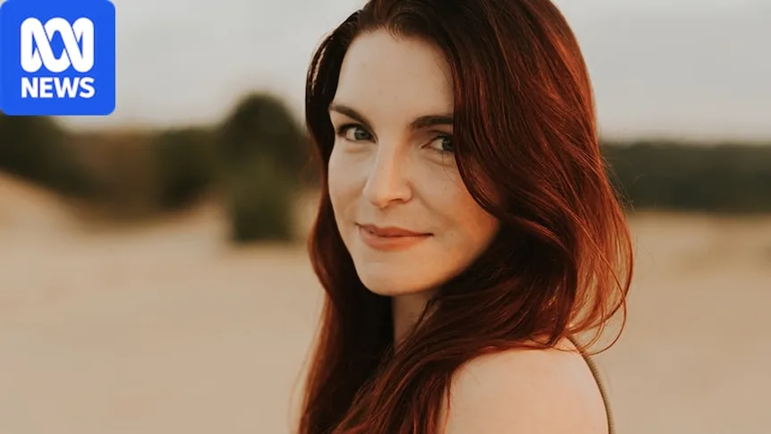 Carissa smiles softly over her shoulder while standing on a beach, wearing a khaki singlet, red hair falling in waves