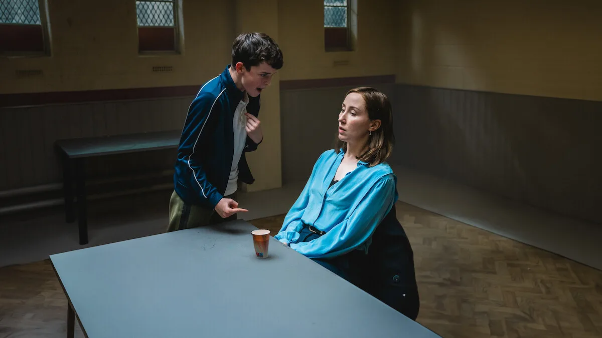 A tense scene shows a young man standing and gesturing intensely at a seated woman in a blue blouse across a table in a dimly lit room with barred windows.