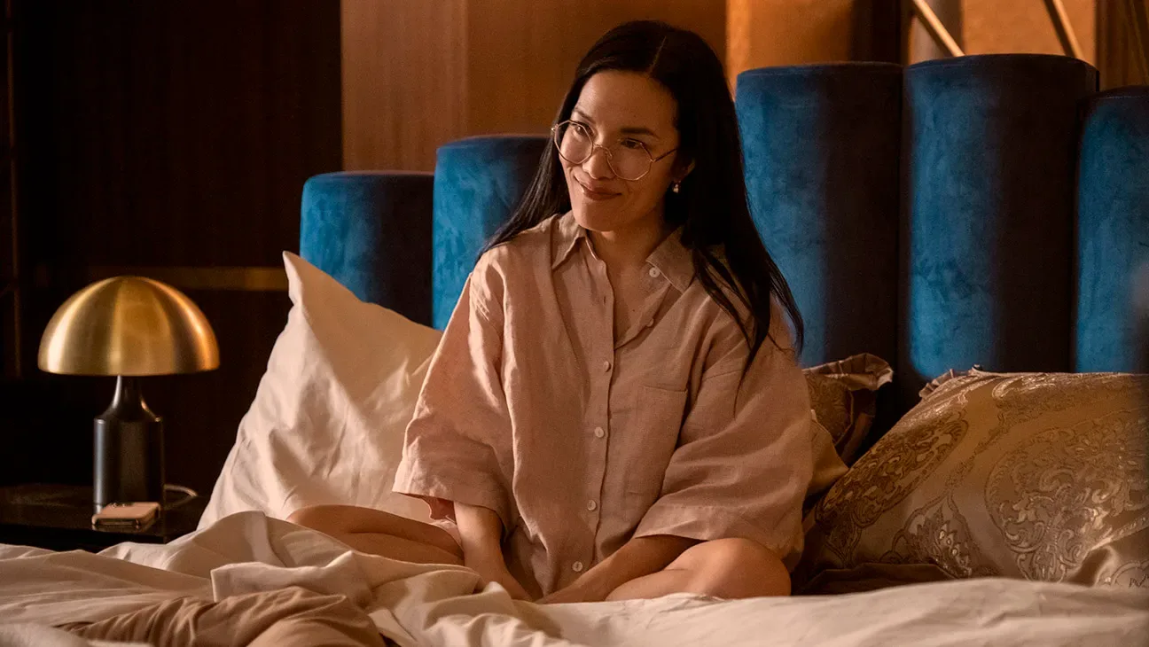 Ali Wong in character in 'Beef,' sitting on a luxurious bed wearing pink pajamas.