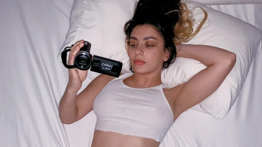 A woman lies on a white bed in a white singlet and underwear, looking at a handycam screen.