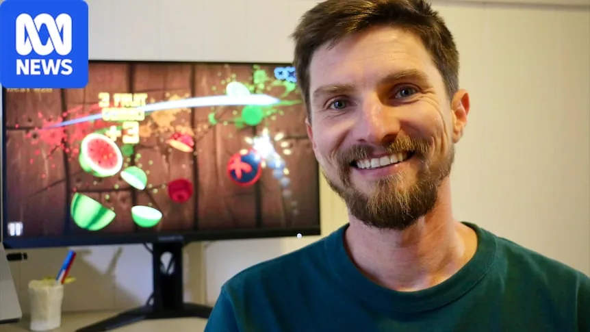Luke Muscat smiling in front of a screen with a still from Fruit Ninja. Fruit being sliced.