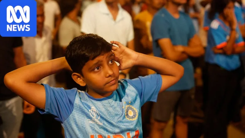 A young Indian boy wearing blue looks sad as he pulls his arms behind him in crowd watching TV