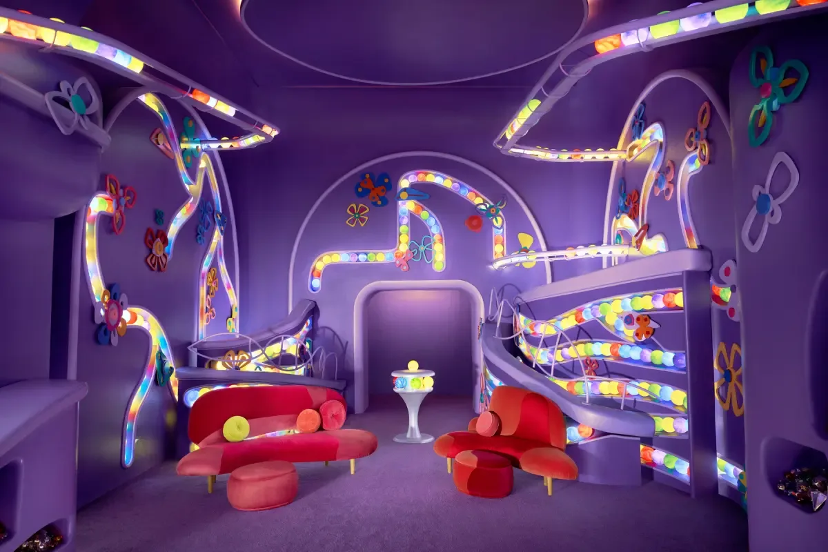 Purple room that has tubes running along the walls filled with green, blue, yellow, purple, and red balls. In the center of the room stand two modern red couches and a pedestal containing the bright…