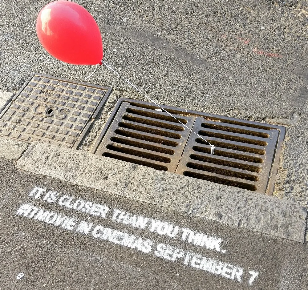 A red balloon tied to a storm drain beside an 'It' street stencil, from a marketing stunt.