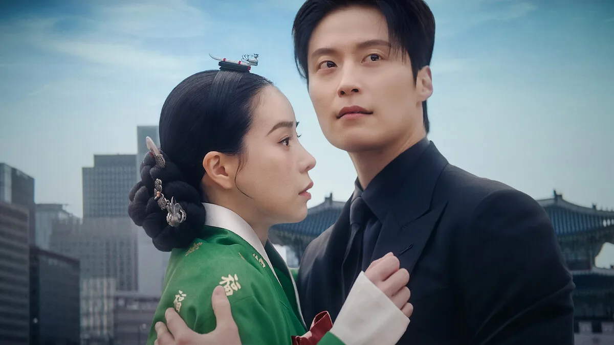 A woman in traditional Korean hanbok and a man in a modern black suit embrace with concern, standing outdoors against a backdrop of city skyscrapers and historical palace architecture under a blue…