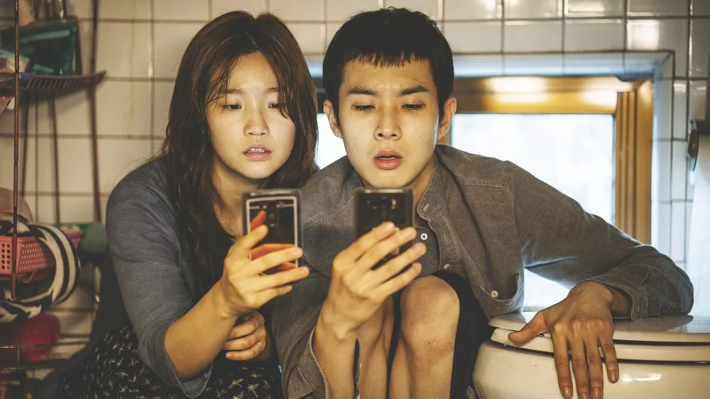 Choi Woo-shik and Park So-dam as the Kim siblings hunting for Wi-Fi in their semi-basement.