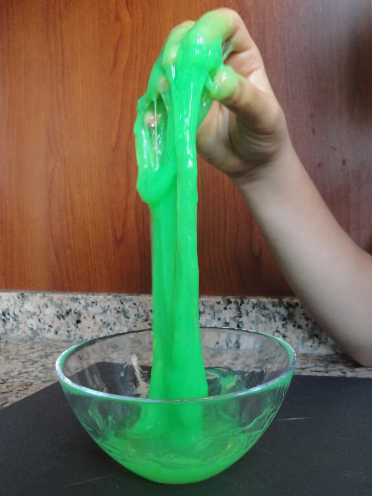 A hand stretching homemade green slime in a glass bowl; CC BY-SA 3.0.