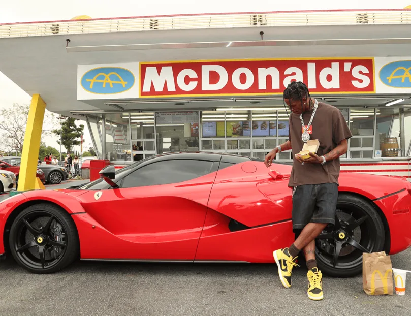 Travis Scott with the meal beside a red Ferrari outside a retro McDonald's.