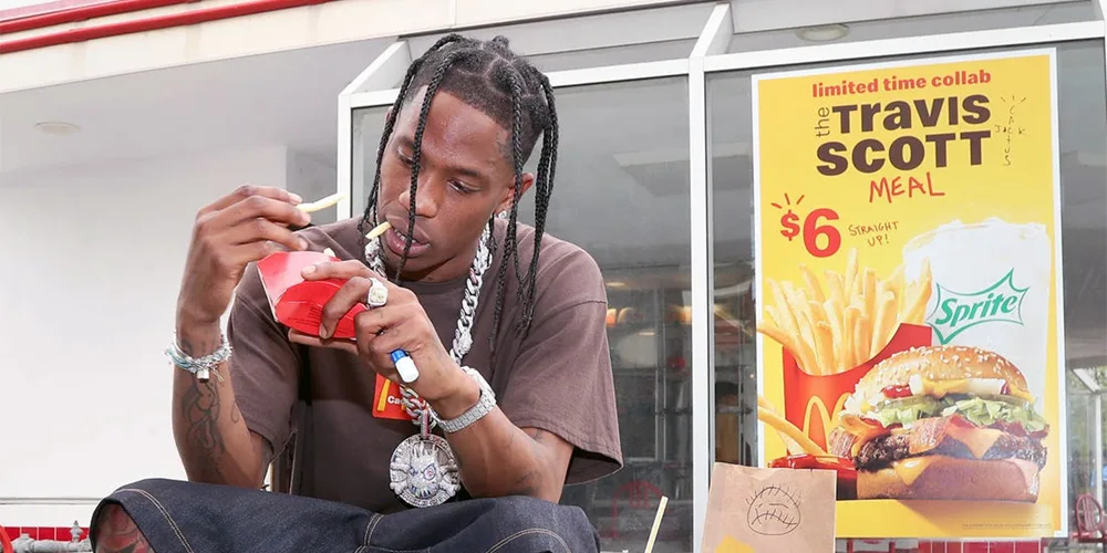 Travis Scott eating fries in front of a Travis Scott Meal poster.